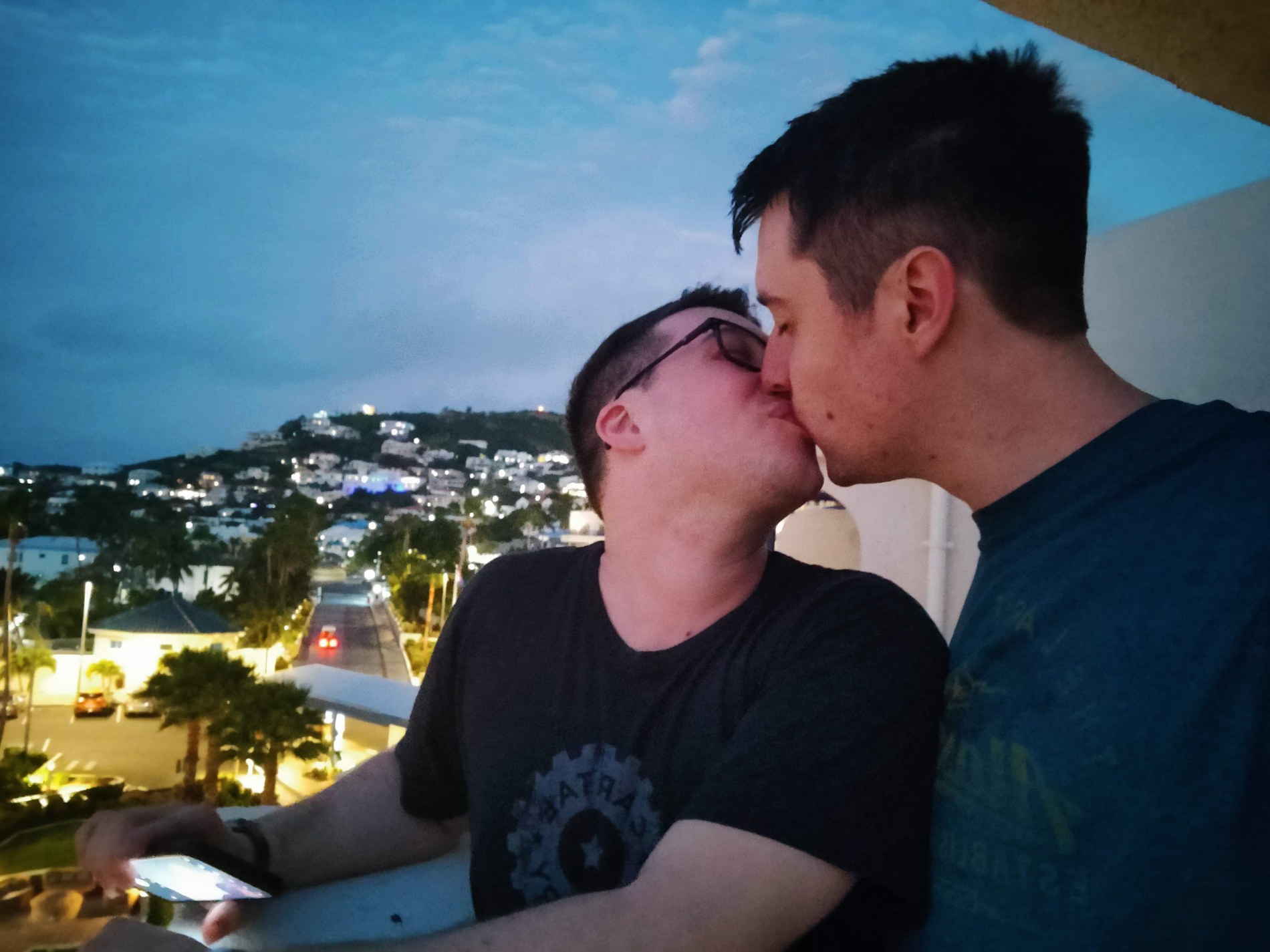 Aaron and Peter sharing a kiss above a hillside town at dusk